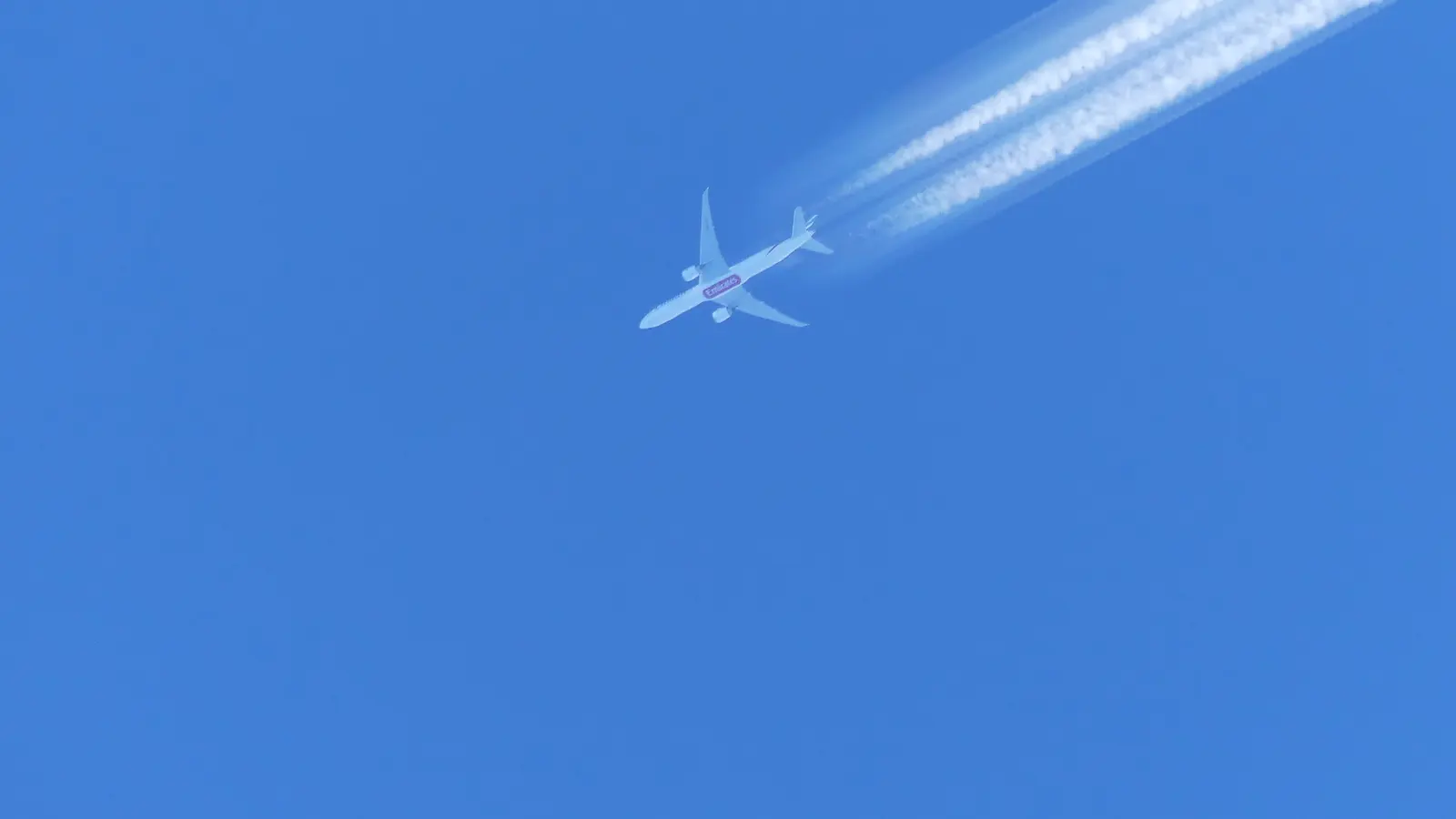 Avião comercial cruza o céu azul deixando duas trilhas brancas de condensação.